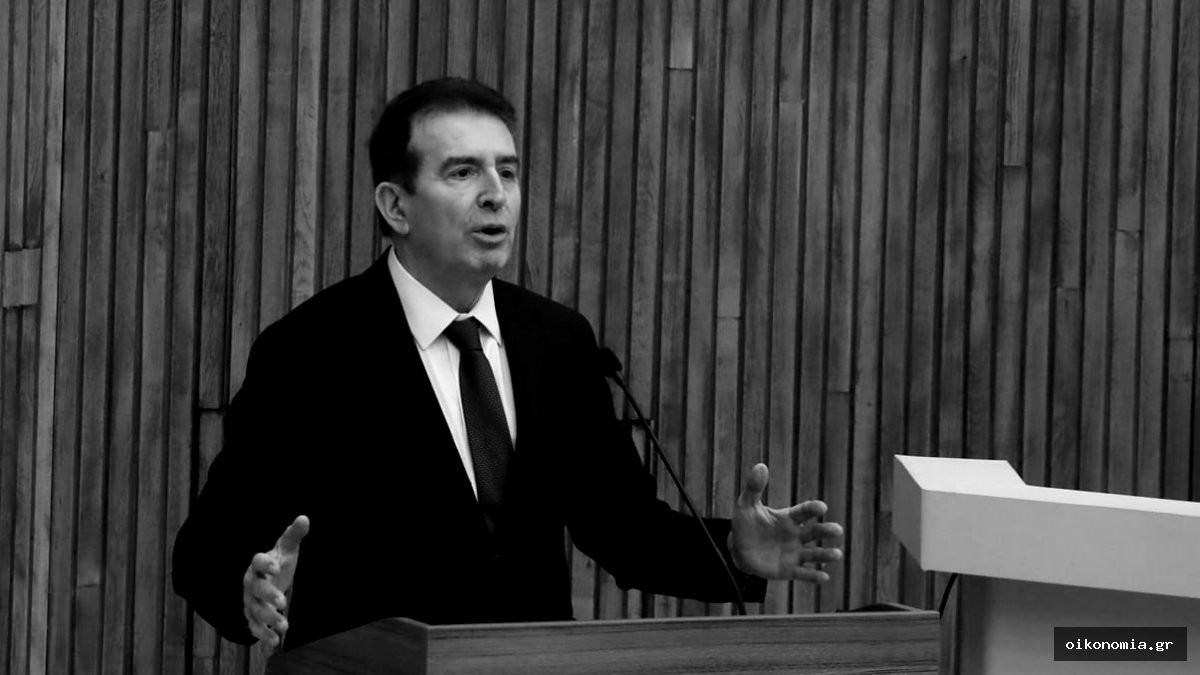 Ο Μιχάλης Χρυσοχοΐδης μιλά σε βήμα φορώντας σκούρο κοστούμι και γραβάτα.