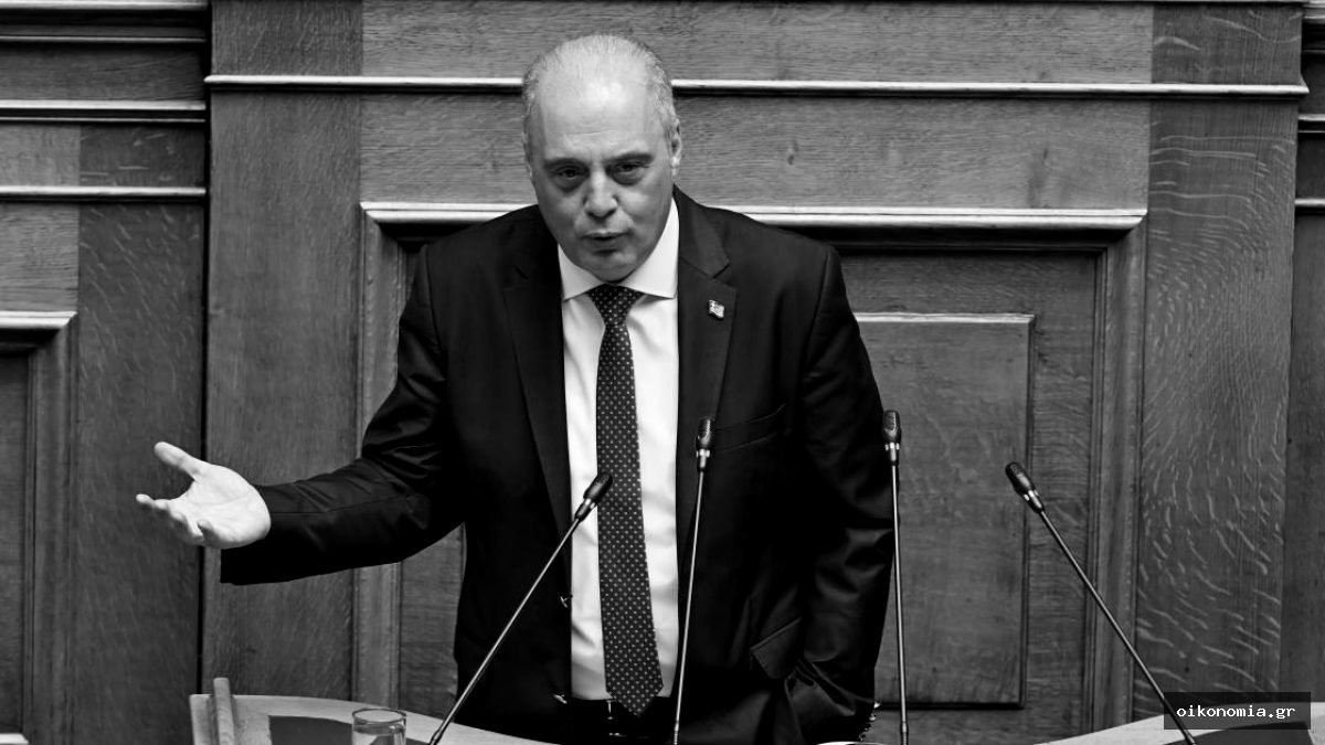 Άνδρας με σκούρο κοστούμι μιλά από το βήμα της Βουλής, μπροστά σε ξύλινη επένδυση.