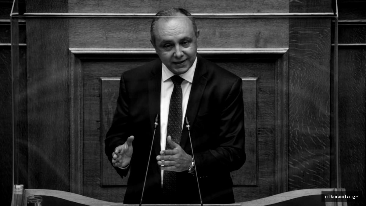 Ο Θεόδωρος Καράογλου μιλά στο βήμα της Βουλής, μπροστά σε ξύλινη επένδυση.