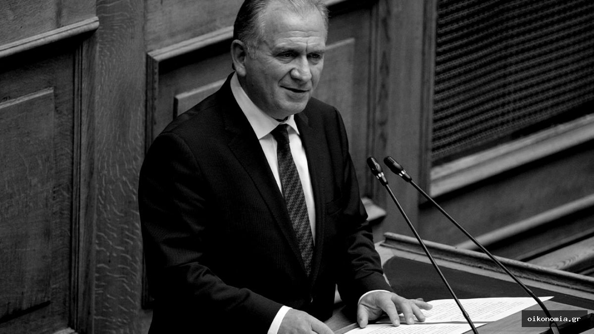 Ο Γιώργος Μουλκιώτης μιλά στο βήμα της Βουλής, μπροστά σε ξύλινη επένδυση και μικρόφωνα.
