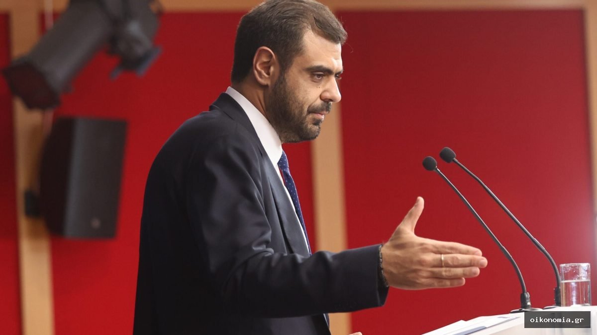 Ο Παύλος Μαρινάκης μιλά σε βήμα με μικρόφωνα, μπροστά σε κόκκινο φόντο.