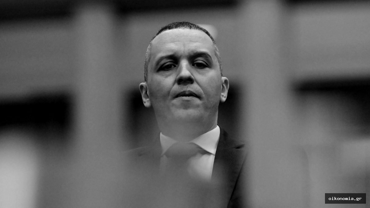 Άνδρας με κοστούμι σε εσωτερικό χώρο, με κάθετα στοιχεία να φαίνονται στο προσκήνιο
