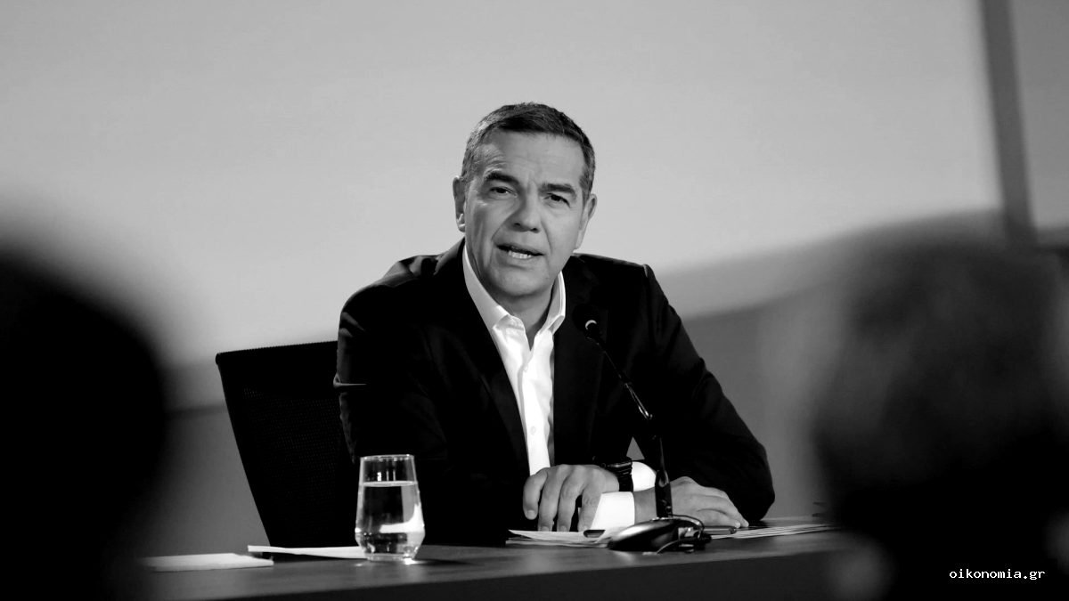 Ο Αλέξης Τσίπρας με σκούρο σακάκι και λευκό πουκάμισο, μπροστά σε μικρόφωνο, ποτήρι νερού και φλιτζάνι.